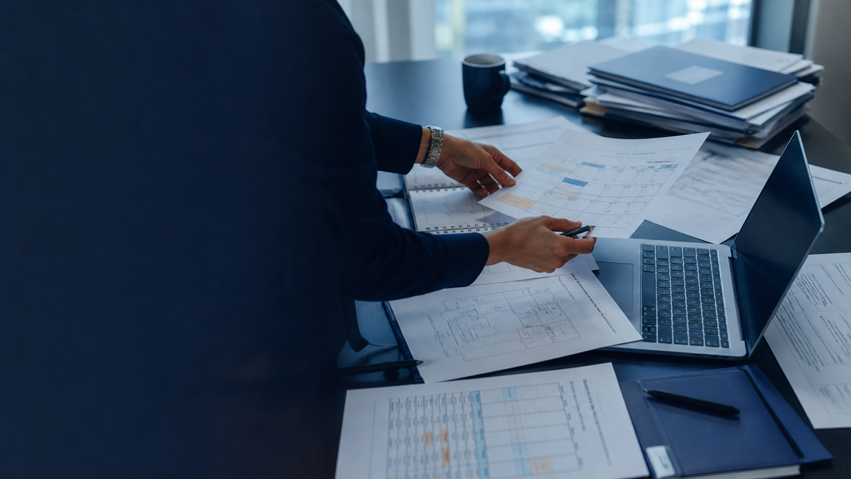 An operations professional reconciling calendars, room plans, and meeting records beside a laptop.