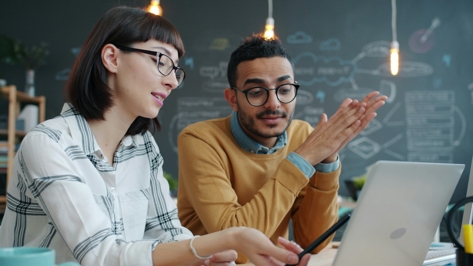 Two colleagues discussing work at a laptop in front of a planning wall.